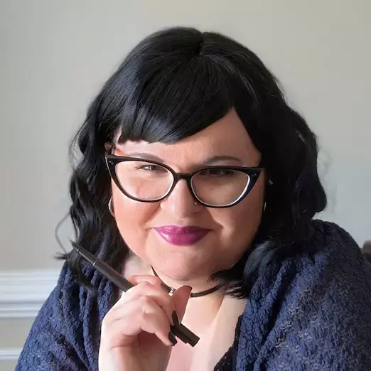 Author E. V. Thorne smiling at the camera, wearing glasses and dark lipstick, holding a black fountain pen.